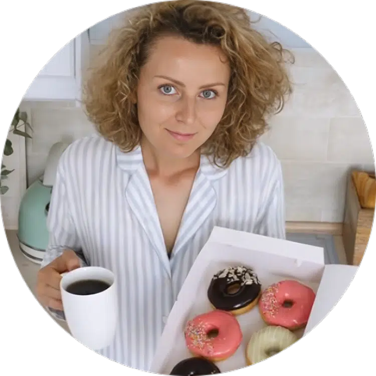 Woman in a striped shirt holding a cup of coffee and a box of assorted colorful donuts in a bright kitchen.