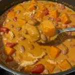 Chicken Pumpkin Chili 5 A bowl of creamy orange-colored chicken pumpkin chili topped with shredded cheese and green onions, served with crusty bread on a wooden table with fall decorations in the background.