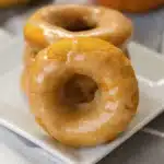 Best Pumpkin Donuts with Maple Glaze 5 Golden brown pumpkin donuts with maple glaze dripping down the sides, arranged on a white plate with fall leaves in the background