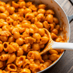 spicy butternut squash pasta in white ceramic bowl topped with dark green crispy sage leaves and white vegan parmesan shreds, fork twirling rigatoni showing creamy sauce coating every piece