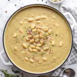 White Bean Soup 8 A steaming bowl of creamy white bean soup with visible chunks of carrots, celery, and white beans, garnished with fresh spinach and grated Parmesan cheese, served with crusty bread on the side.