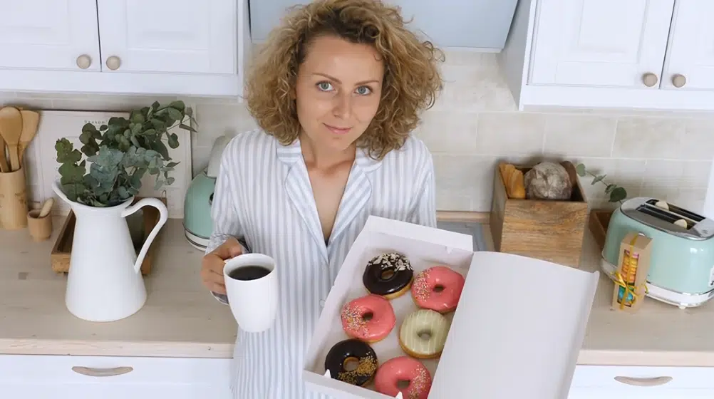 About me 1 Woman in a striped shirt holding a cup of coffee and a box of assorted colorful donuts in a bright kitchen.