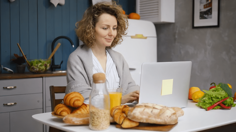 inez in a striped shirt and shawl working on a laptop at a kitchen table with fresh bread, croissants, orange juice, and cereal