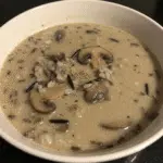 Wild Rice and Mushroom Soup 6 Steaming bowl of creamy wild rice and mushroom soup with golden brown mushrooms floating on top, served with crusty bread on a wooden kitchen table