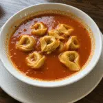 A bowl of creamy tomato tortellini soup garnished with fresh basil leaves, served alongside crusty bread, photographed from above on a rustic wooden table