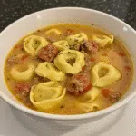 Italian Sausage Tortellini Soup 6 A steaming bowl of creamy Italian sausage tortellini soup with visible cheese tortellini, spinach leaves, and herbs, served in a rustic white bowl with crusty bread on the side