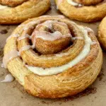 Golden, flaky puff pastry cinnamon rolls arranged on a white plate, drizzled with vanilla glaze and topped with chopped toasted pecans, photographed from above on a marble countertop.