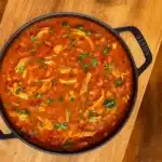 A steaming bowl of poblano chicken tortilla soup garnished with avocado slices, shredded cheese, and tortilla strips, with lime wedges on the side