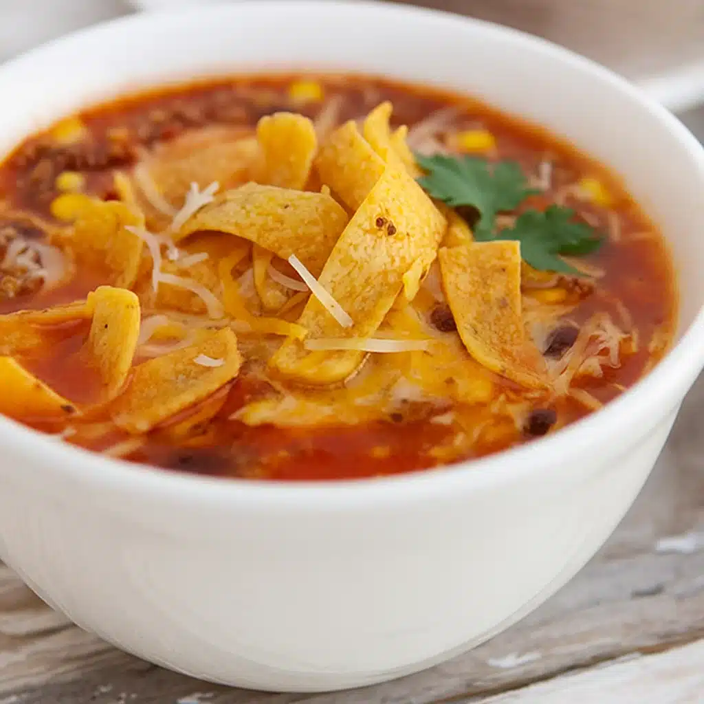 Taco Soup Recipe 4 Steaming bowl of hearty taco soup topped with shredded cheese, diced avocado, and tortilla strips on rustic wooden table