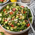 A fresh chickpea feta avocado salad in a white bowl with creamy avocado chunks, chickpeas, crumbled feta cheese, and fresh cilantro, garnished with lime wedges