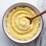 A white bowl filled with creamy garlic mashed potatoes topped with a pat of melting butter and fresh chopped parsley, with a silver spoon beside it