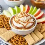 Layered caramel apple cheesecake dip in a glass pie dish showing distinct layers of graham cracker crumbs, creamy cheesecake filling, caramel sauce, whipped topping, and chopped peanuts, surrounded by fresh apple slices and graham crackers for dipping