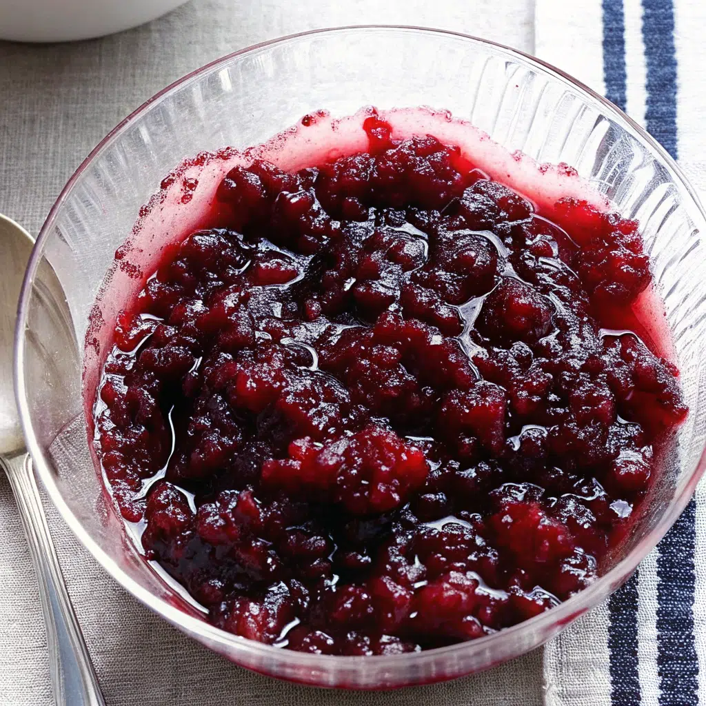 A bowl of glossy, ruby-red homemade spiced cranberry sauce garnished with orange zest and a cinnamon stick, served in a white ceramic bowl on a rustic wooden table