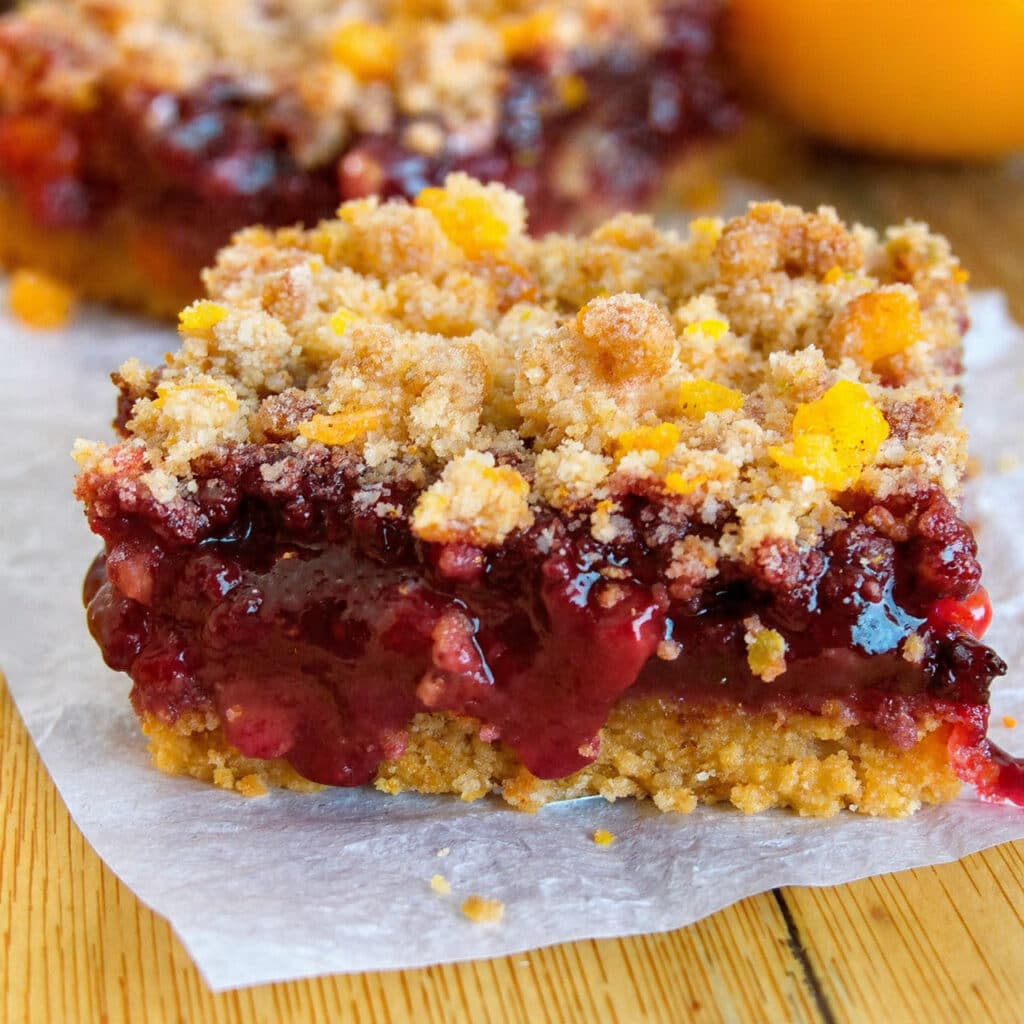 Thick square cranberry orange crumb bars with chunky golden oat topping and bright red cranberry filling visible on cut sides, three bars stacked on white plate