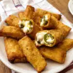 Golden-brown crispy mini jalapeño popper egg rolls arranged on a white plate with a small bowl of ranch dressing for dipping, with one egg roll cut open to show the creamy cheese and bacon filling inside.
