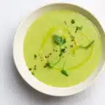 A bowl of vibrant bright green cream spring vegetable soup topped with a swirl of Greek yogurt and fresh chives, served with crusty bread on a white table.
