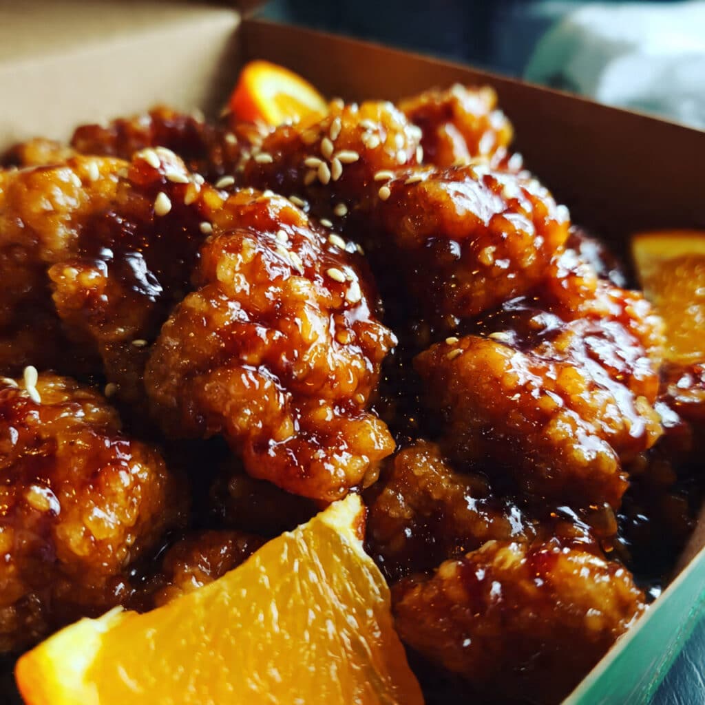 Glazed orange chicken pieces on a white plate over fluffy white rice, garnished with sliced green onions and sesame seeds, with a spoon drizzling extra orange BBQ sauce over the top