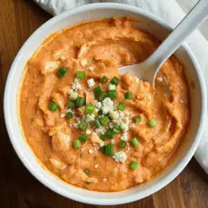 Creamy buffalo chicken dip in a small casserole dish topped with melted golden cheddar cheese and fresh green onions, served with tortilla chips and celery sticks
