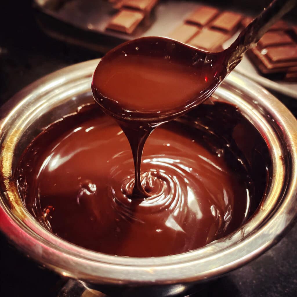 chocolate ganache being poured from a small bowl over a chocolate cake