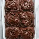chocolate cinnamon rolls arranged in a baking dish, topped with glossy chocolate cream cheese glaze, with visible chocolate swirls throughout the soft dough