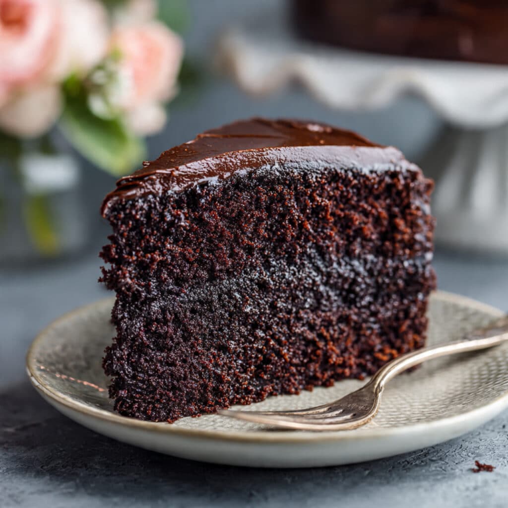 Slice of rich dark chocolate layer cake with chocolate frosting on a white plate, showing moist, tender crumb texture