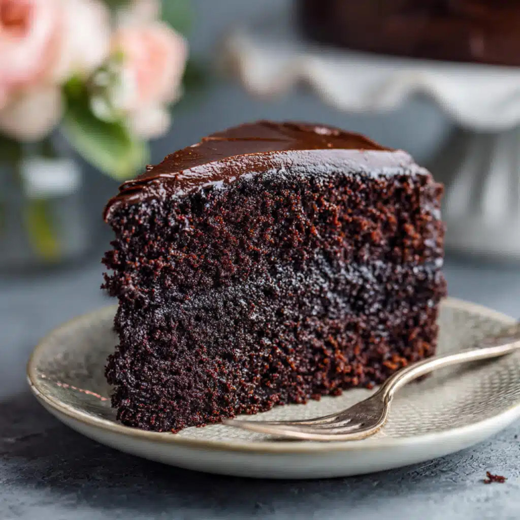 Slice of rich dark chocolate layer cake with chocolate frosting on a white plate, showing moist, tender crumb texture