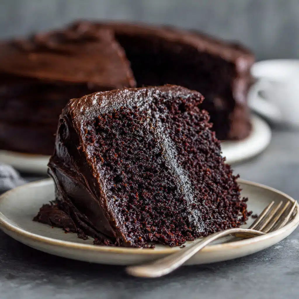 Slice of rich dark chocolate layer cake with chocolate frosting on a white plate, showing moist, tender crumb texture