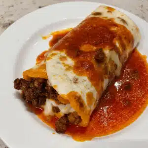 Golden baked wet burritos smothered in red sauce and melted cheese in a baking dish, topped with sour cream and fresh cilantro