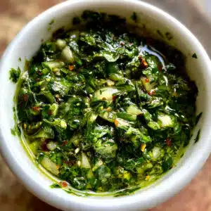 Bright green chimichurri sauce in a small glass bowl with a spoon, surrounded by fresh parsley, garlic cloves, and olive oil on a wooden table