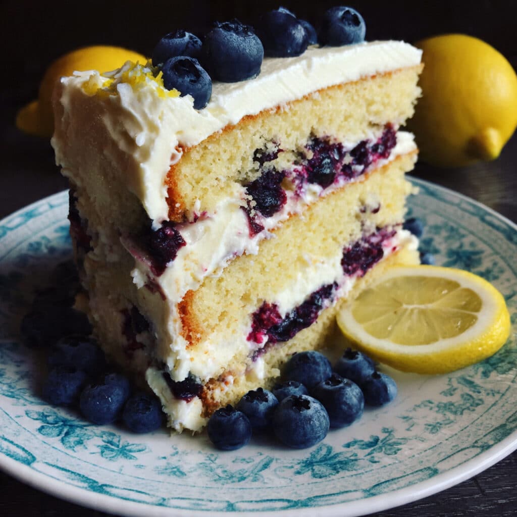 Three-layer lemon blueberry cake with mascarpone frosting, fresh blueberries studded throughout, topped with fresh berries and lemon slices on a white cake stand