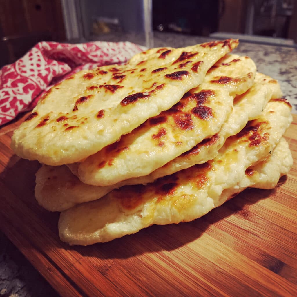 Golden-brown homemade Greek yogurt naan bread stacked on a white plate, brushed with melted butter and garnished with fresh herbs