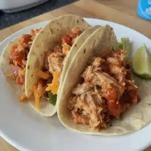 Shredded crockpot chicken tacos on soft flour tortillas topped with fresh lettuce, diced tomatoes, shredded cheese, sour cream, and cilantro on a colorful plate