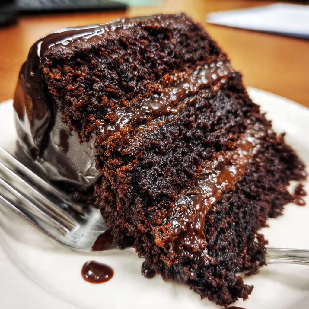 Three-layer chocolate fudge cake with rich dark chocolate frosting on a white cake stand, showing moist cake layers and smooth glossy icing