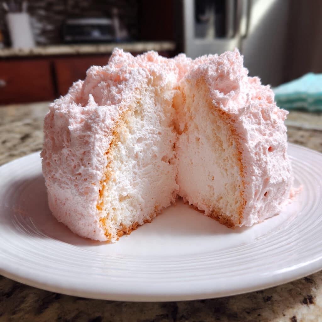 A slice of light, fluffy pink angel food cake on a white plate with fresh strawberries and whipped cream, showing the airy, tender crumb structure