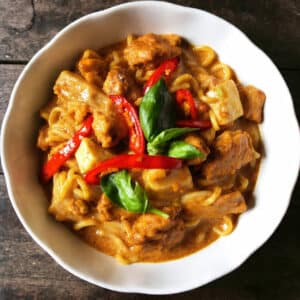 Thai Drunken Noodles (Pad Kee Mao) in a white bowl, showing wide rice noodles coated in dark sauce with pieces of chicken, fresh Thai basil leaves, sliced onions, and red chilies
