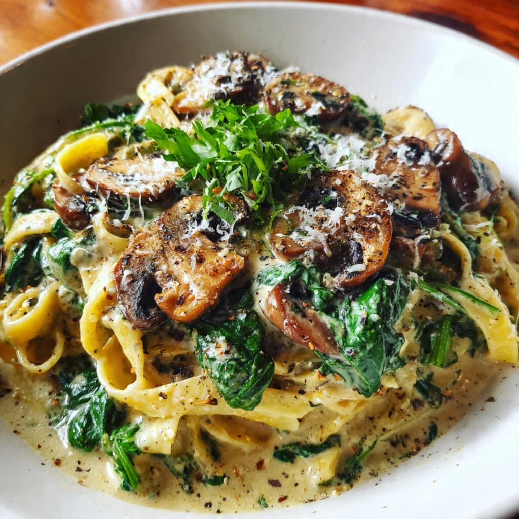 Creamy spinach mushroom pasta in a white bowl, garnished with freshly grated Parmesan cheese and black pepper, with a fork twirling pasta