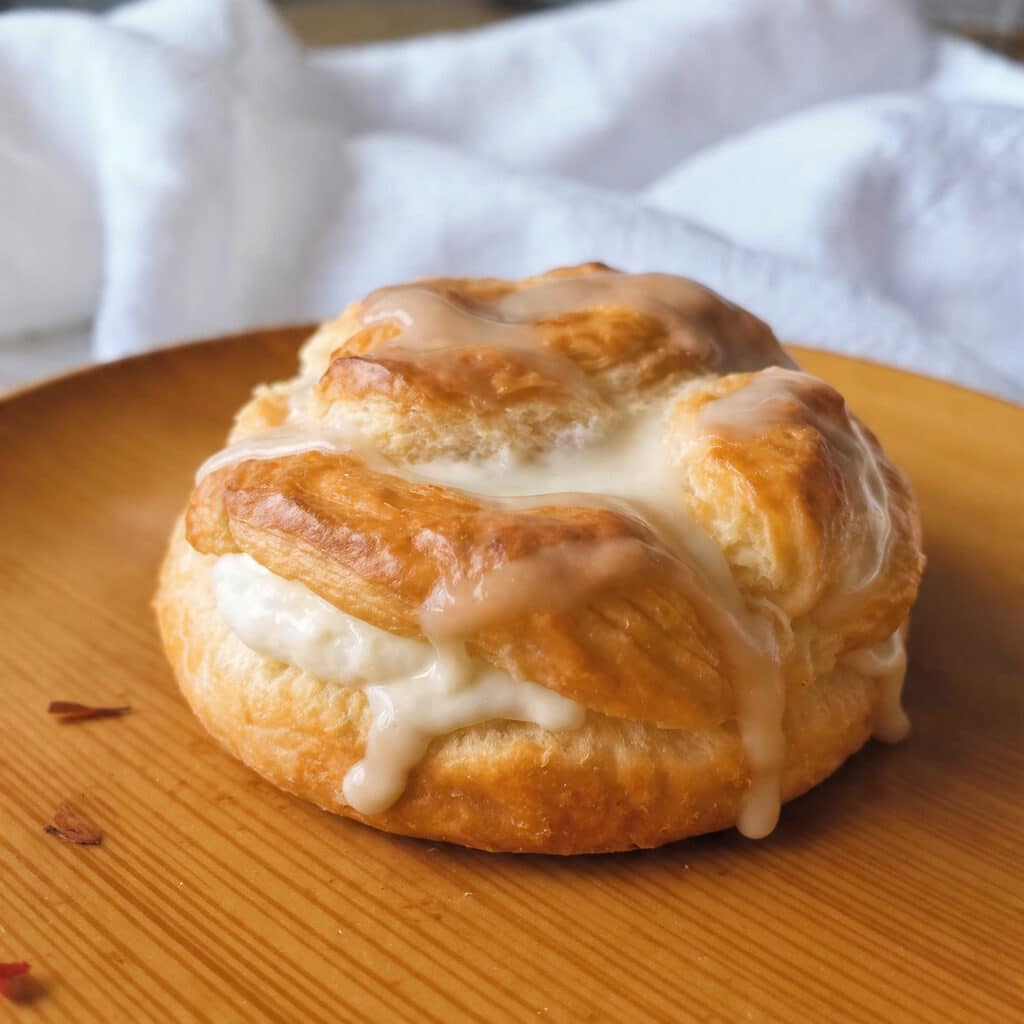 Golden brown cheese danish pastries with cream cheese filling and white glaze drizzled on top, arranged on a white plate with coffee in the background