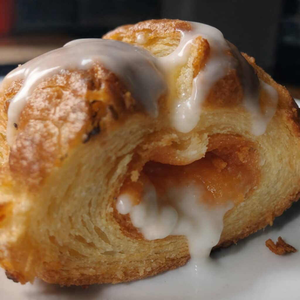 Golden brown cheese danish pastries with cream cheese filling and white glaze drizzled on top, arranged on a white plate with coffee in the background