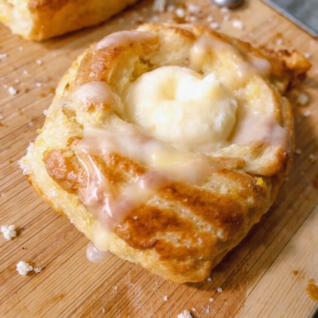 Golden brown cheese danish pastries with cream cheese filling and white glaze drizzled on top, arranged on a white plate with coffee in the background