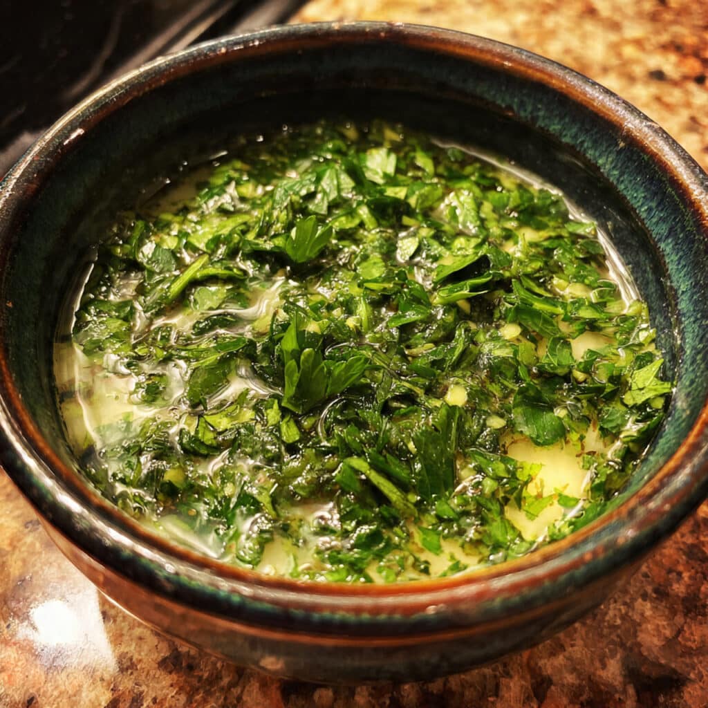 Bright green chimichurri sauce in a small glass bowl with a spoon, surrounded by fresh parsley, garlic cloves, and olive oil on a wooden table