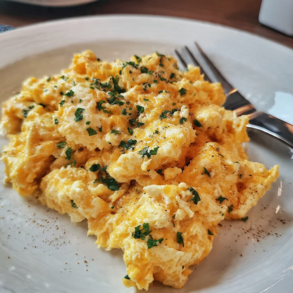 Creamy scrambled eggs with cottage cheese on a white plate, softly folded with visible curds, garnished with fresh black pepper and chives, with a fork on the side and buttered toast in the background