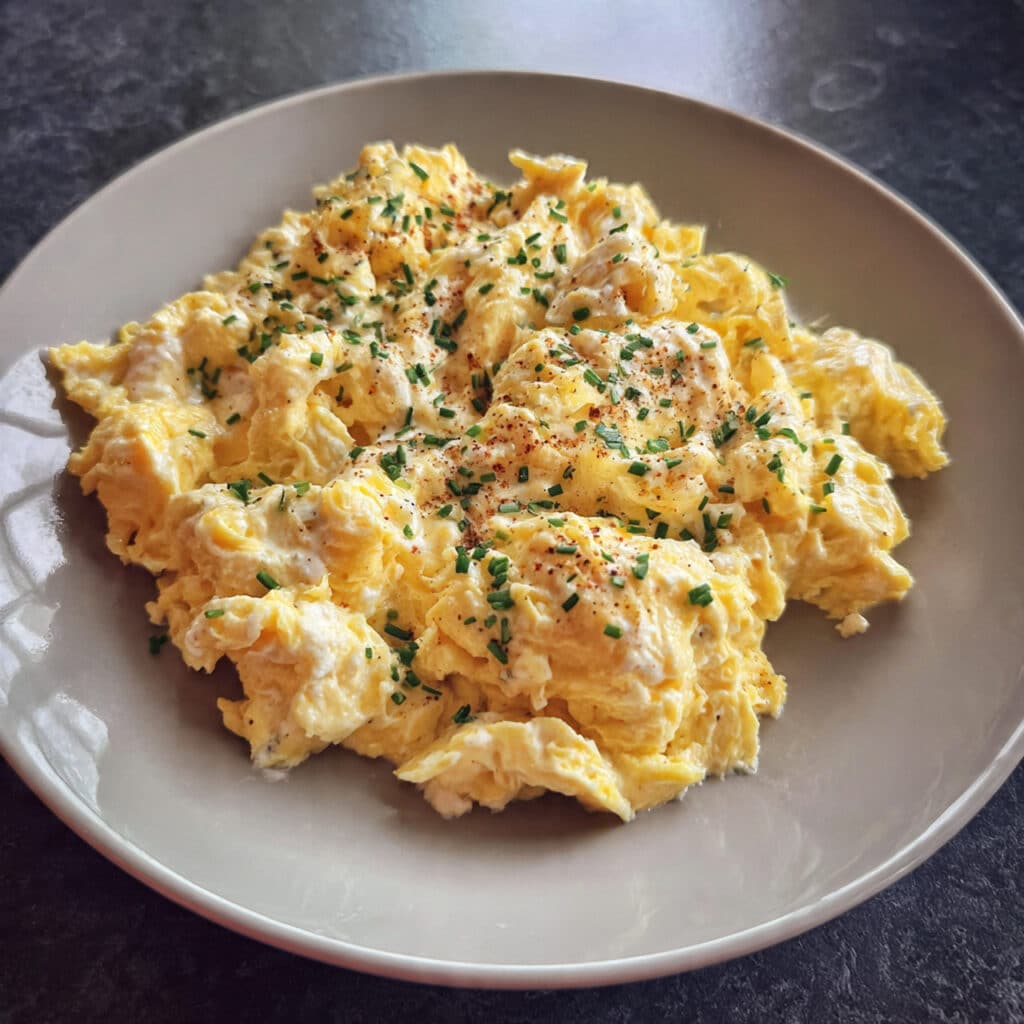 Creamy scrambled eggs with cottage cheese on a white plate, softly folded with visible curds, garnished with fresh black pepper and chives, with a fork on the side and buttered toast in the background