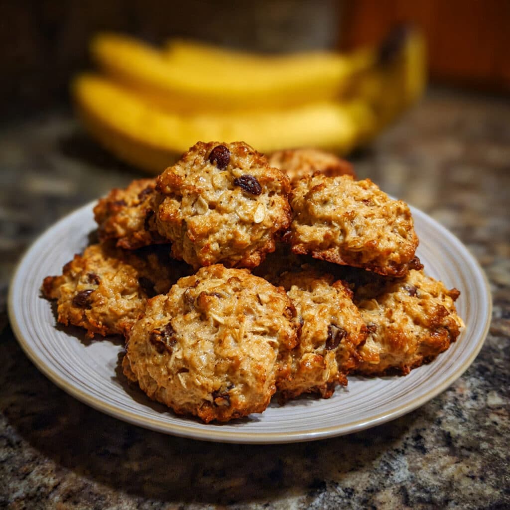 Golden brown banana oatmeal cookies arranged on a white plate with chocolate chips visible, soft and chewy texture, rustic kitchen background