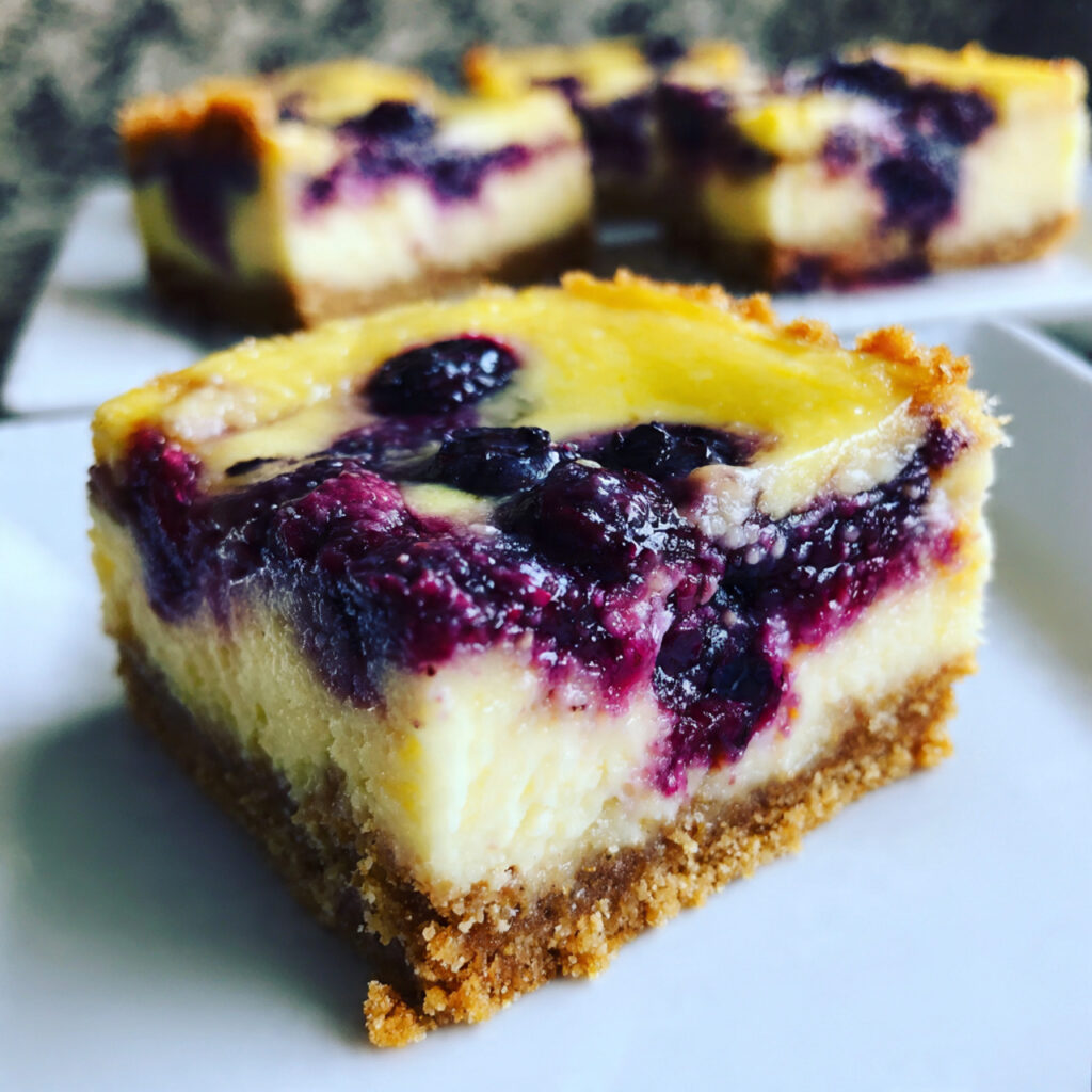 Square blueberry lemon cheesecake bars on a white plate showing creamy yellow cheesecake with purple blueberry swirls and golden graham cracker crust