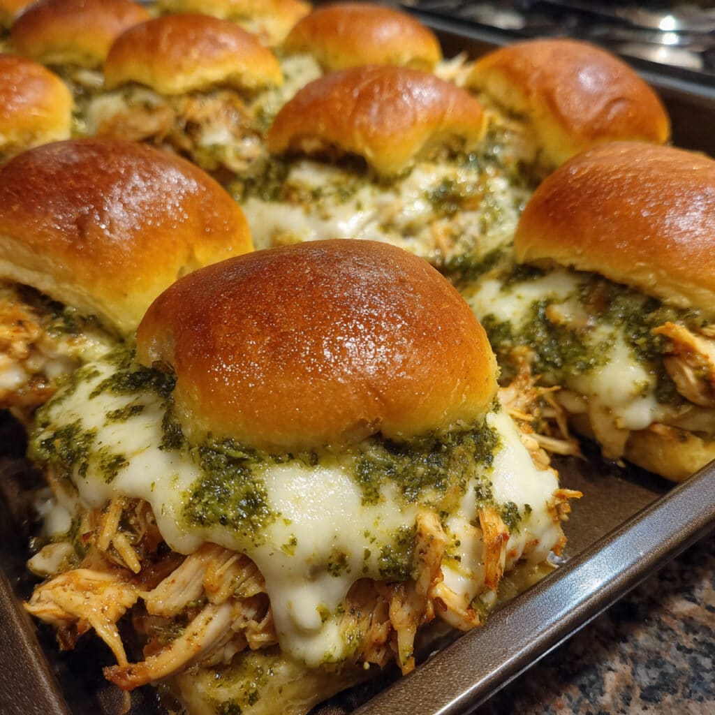 Golden-brown cheesy garlic pesto chicken sliders in a baking dish, showing melted mozzarella and herb-topped slider buns