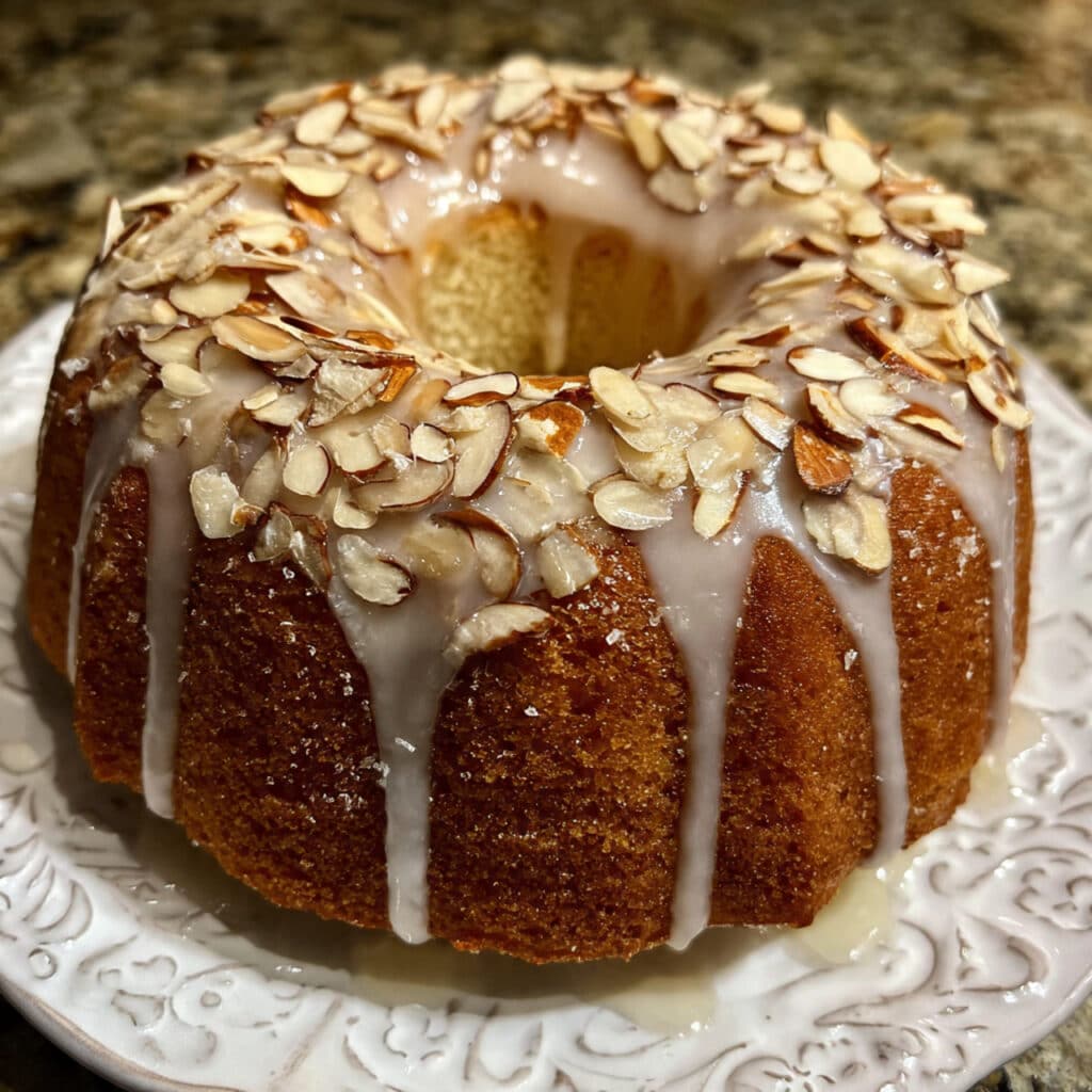 golden brown almond bundt cake on a white cake stand, drizzled with white almond glaze and topped with toasted sliced almonds, with one slice removed showing the moist, tender crumb