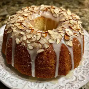 golden brown almond bundt cake on a white cake stand, drizzled with white almond glaze and topped with toasted sliced almonds, with one slice removed showing the moist, tender crumb