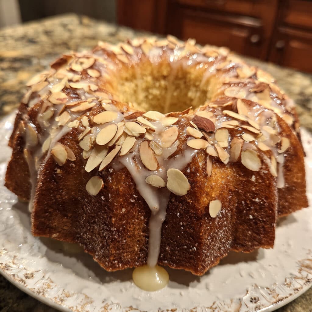 golden brown almond bundt cake on a white cake stand, drizzled with white almond glaze and topped with toasted sliced almonds, with one slice removed showing the moist, tender crumb