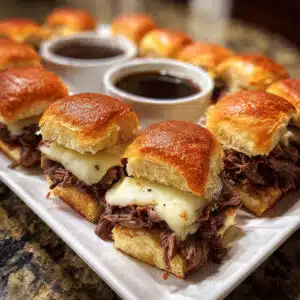Golden brown French dip sliders on a white platter with melted cheese and tender beef, served alongside small bowls of rich au jus dipping sauce
