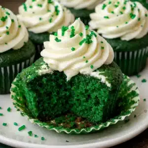 Vibrant green velvet cupcakes topped with swirls of white cream cheese frosting and festive shamrock sprinkles on a white serving plate